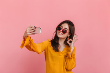 Female influencer in a yellow top taking a selfie with a pink background, symbolizing social media content creation.